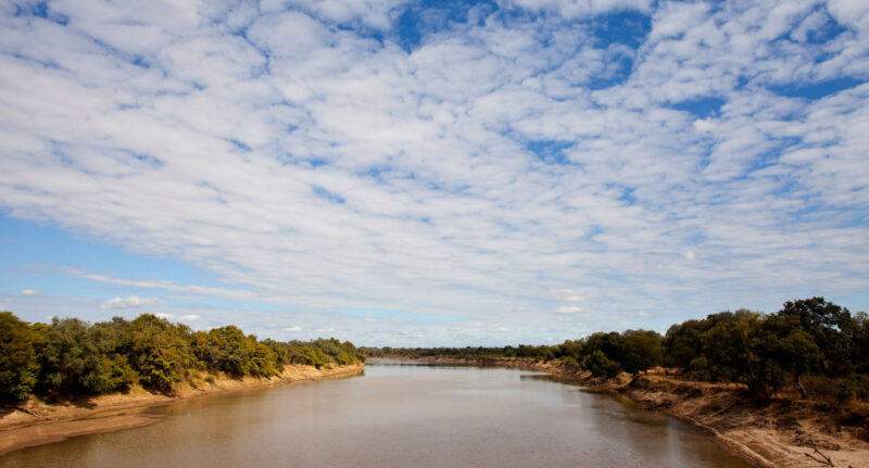 Wide landscape view of a calm river between tree-lined banks under a bright sky with many small white clouds.