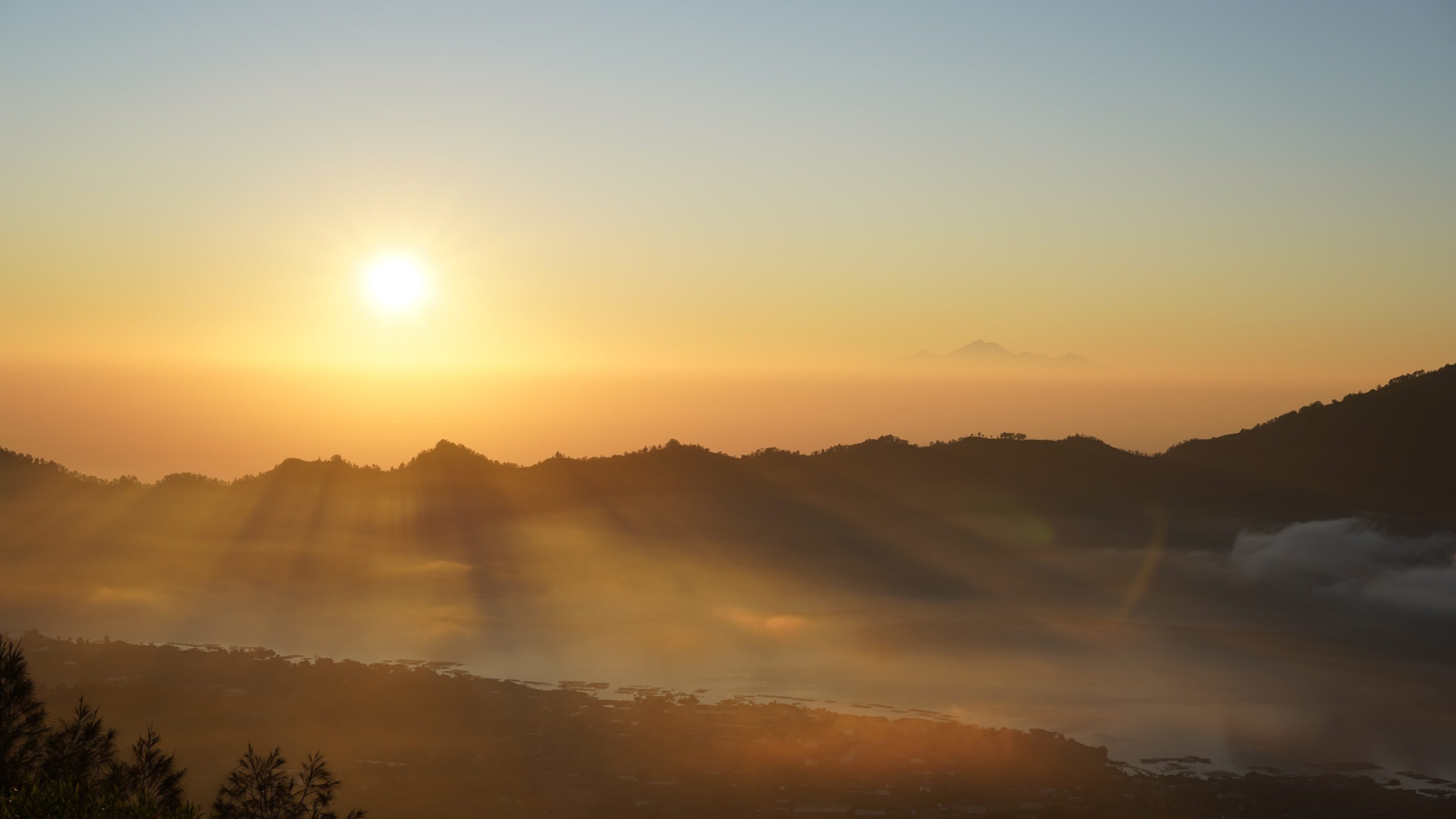 Sun rays breaking through clouds over a dark mountain range at sunrise with mist filling the valley below.