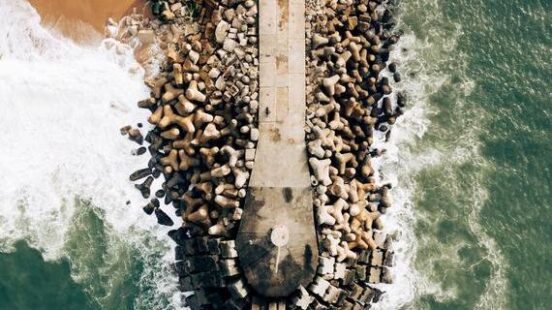 Top-down view of a concrete pier surrounded by interlocking stone blocks separating the beach from green ocean waves.