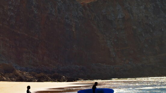 Silhouetted people and a surfer with a blue board walk along a sandy beach near high, rugged cliffs.