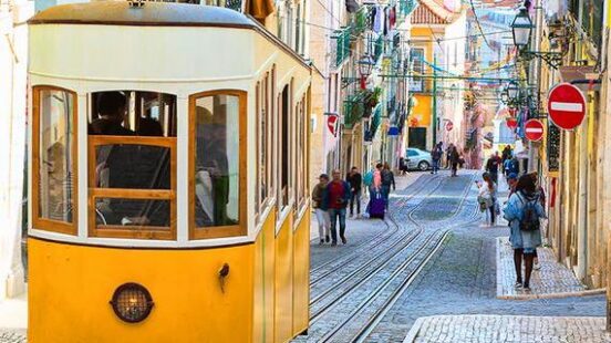 A yellow and white funicular tram travels down a steep, narrow city street with colorful buildings and pedestrians.