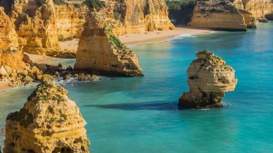 Tall, rugged rock formations stand in clear turquoise ocean water near a sandy beach and limestone cliffs.