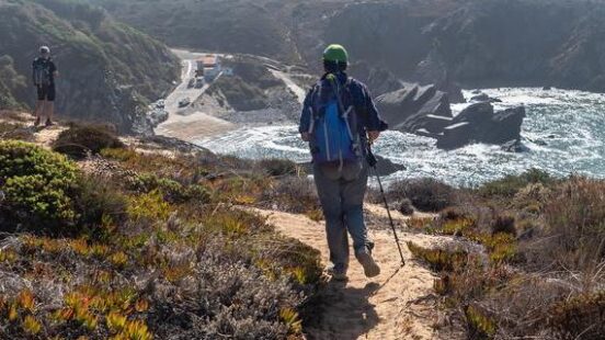A person with a blue backpack hikes a sandy trail on a cliffside overlooking the ocean and rocky shoreline.