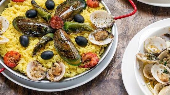 Multiple plates of seafood including yellow rice with mussels, shrimp in garlic oil, and steamed clams.