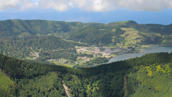 A large green lake sits inside a deep volcanic crater surrounded by steep, forest-covered hills and white clouds.