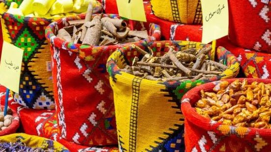 Colorful woven bags filled with bulk spices and herbs including cinnamon sticks and dried flowers at a market.