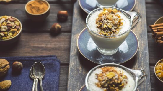 Cups of Mahalabia pudding garnished with nuts and dried fruit served on a dark wood table with cinnamon sticks.