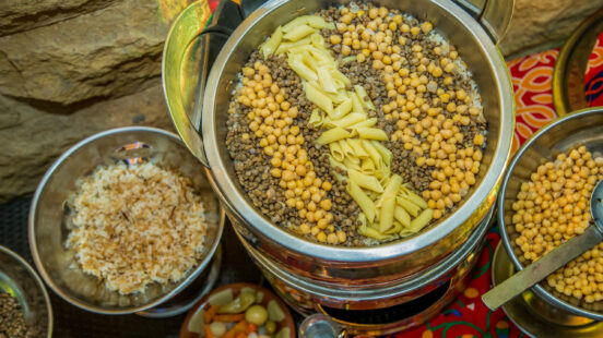 Overhead view of a large catering pot containing koshary ingredients arranged in neat rows of pasta and legumes.