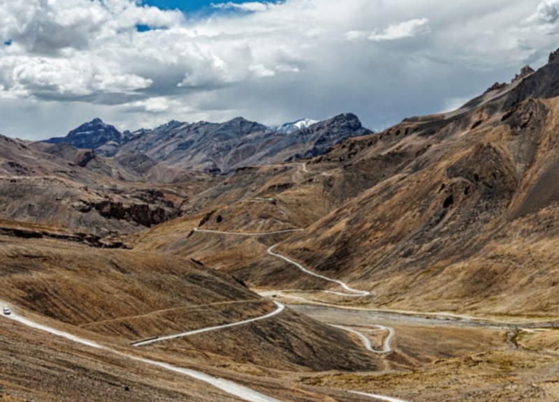 A mountain road snakes through the rugged, high-altitude desert terrain of Ladakh, under a dramatic sky.