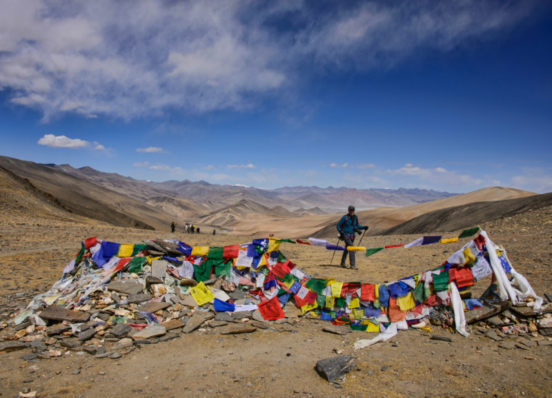 A trekker pauses by vibrant prayer flags fluttering across a high-altitude mountain pass in Ladakh.