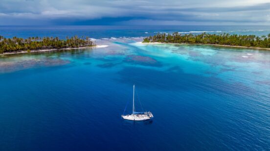 Aerial view of a white sailboat anchored in blue water between two tropical islands with palm trees.