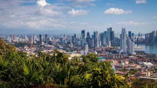 Wide view of a dense city skyline with tall modern skyscrapers bordering a blue ocean bay under a bright sky.