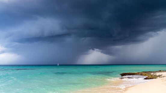 Large dark storm clouds over a turquoise ocean with a sandy beach in the foreground and a sailboat in the distance.