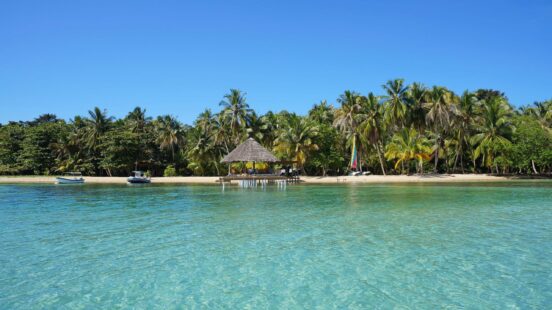 A thatched roof hut on a pier over clear turquoise water with a palm tree lined beach in the background.