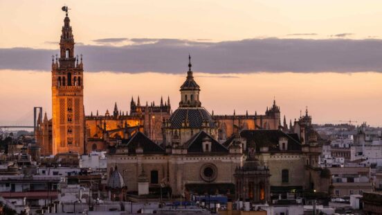 A cityscape of Seville at twilight featuring the prominent Giralda tower and ornate cathedral domes against a pink sky.