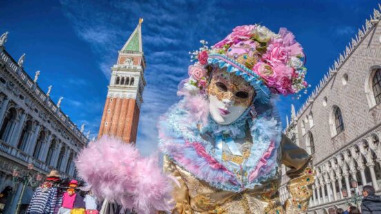 A person in a blue and pink ornate mask and costume stands in St. Markx27;s Square with the bell tower behind them.