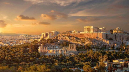 The ancient Acropolis of Athens sits atop a rocky hill overlooking the city under a warm sunset sky.