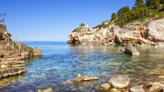 Clear blue water in a rocky coastal inlet with sunlit cliffs and a stone walkway.