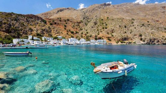 A white boat floats on transparent turquoise water in front of a seaside village and mountain.