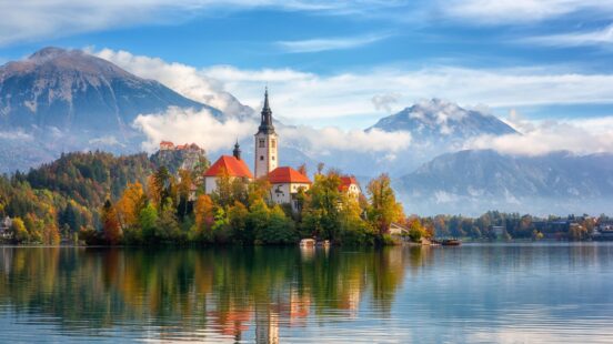 A white church with a tall steeple on a small island in the middle of a calm lake.