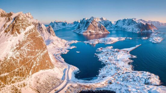 Aerial view of snow-capped mountains surrounding a small coastal town and blue fjord.