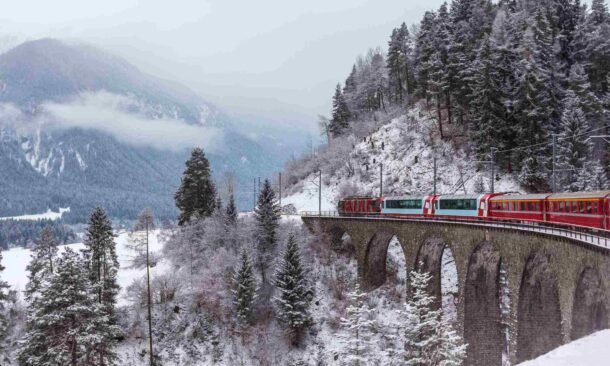A red train traveling over a stone bridge in a winter mountain setting with snow-covered trees.