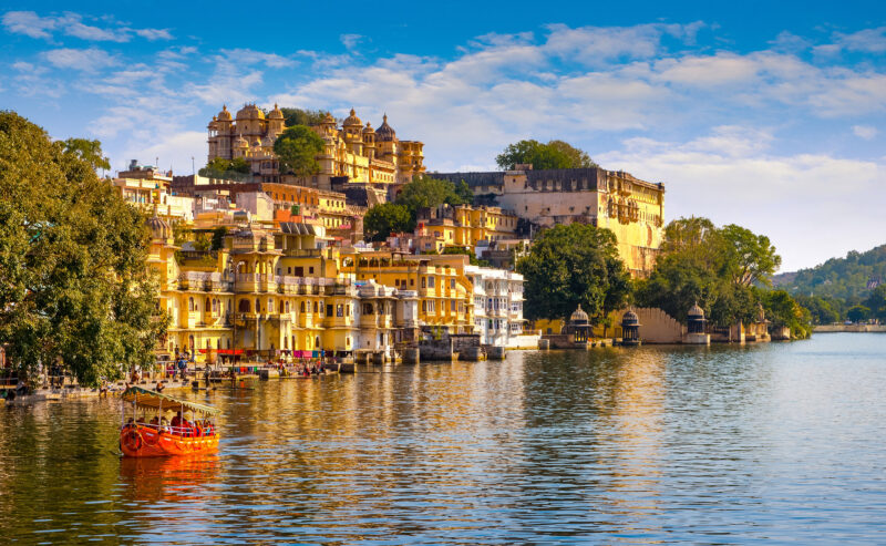 A large yellow palace complex sits on the edge of a lake with a small orange tour boat on the water.
