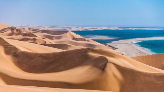 High angle view of rolling sand dunes bordering a blue ocean under a clear sky.