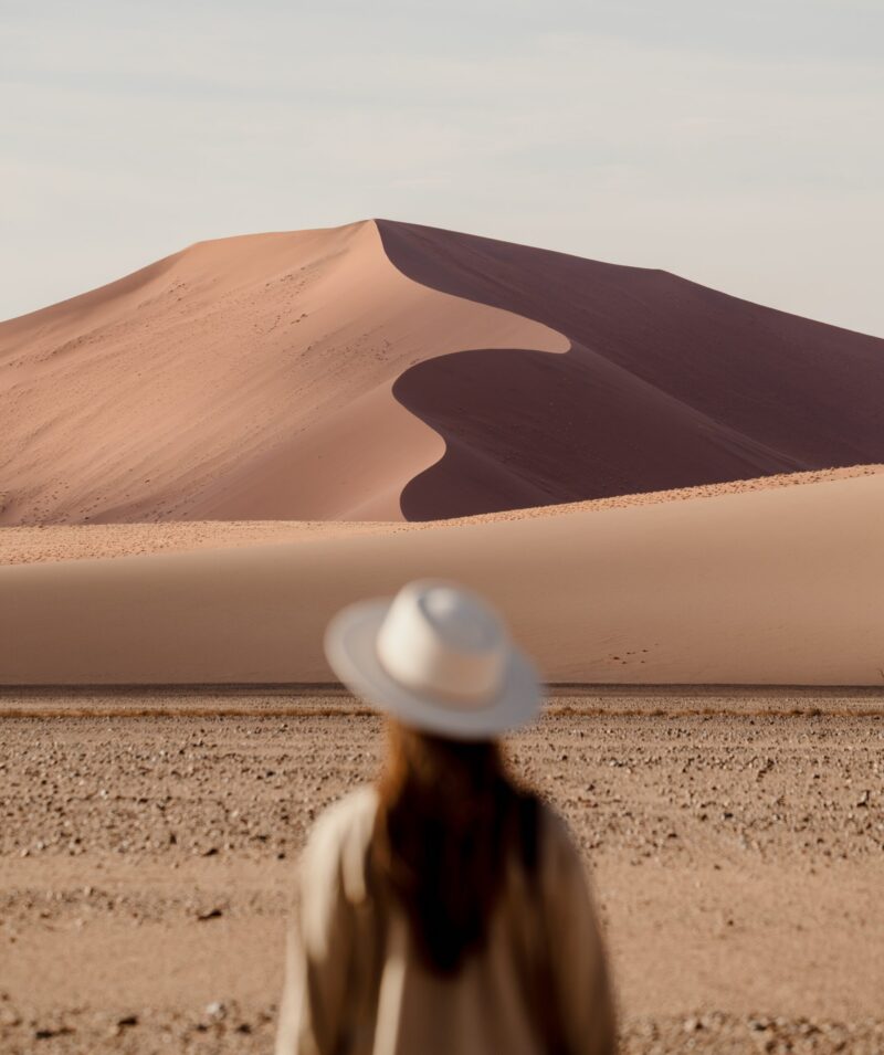Over-the-shoulder view of a person in a hat looking at a tall orange sand dune with a curved crest.