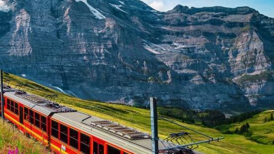 A red and yellow mountain train climbs a green meadow with purple flowers below a massive snow-capped peak.