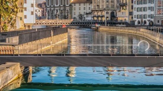 Water cascades over a weir on a river in Lucerne with historic buildings and the Jesuit Church in the background.