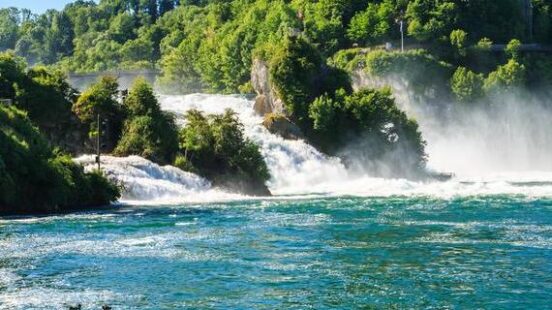 The wide, powerful Rhine Falls rushing over rocks, with a white castle nestled in green trees on the cliff above.
