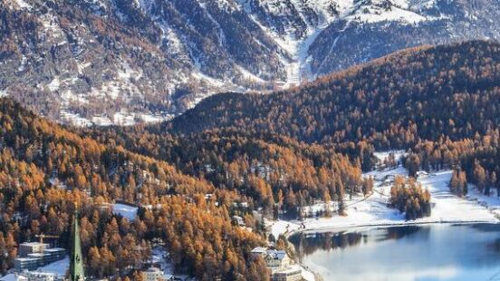 St. Moritz town and lake with autumn trees and heavy snow-capped mountains in the background.