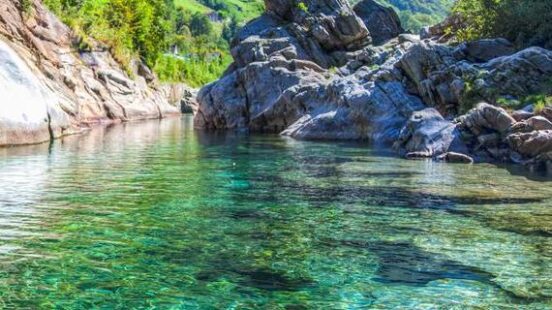A double-arched stone bridge spans the shallow, crystal-clear emerald-green river in a rocky, green valley.