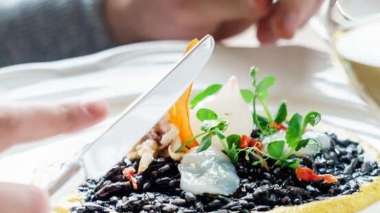 A personx27;s hands using a knife and fork to eat black risotto topped with seafood and microgreens on a white plate.