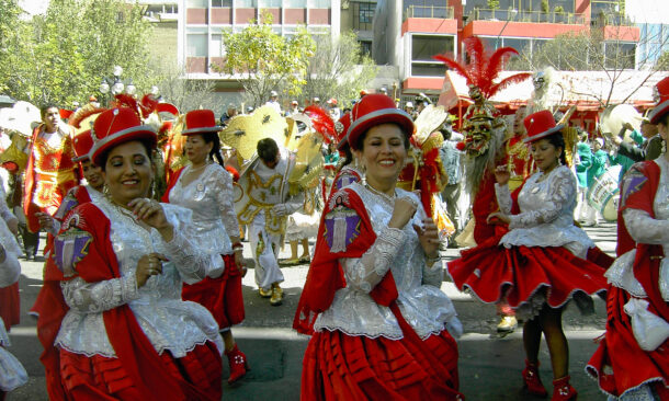 Women in traditional red and white costumes and hats dance in a street parade with buildings in the background.