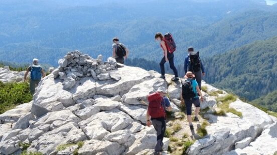 Hikers with backpacks climbing over large white rocks on a mountain peak with distant hazy blue ridges.