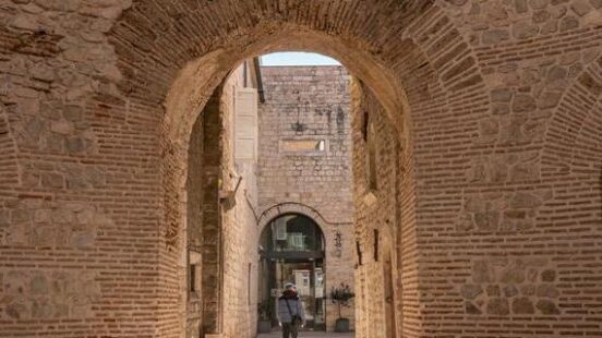 A person walking through a large ancient stone archway into a sunlit courtyard in an old city.