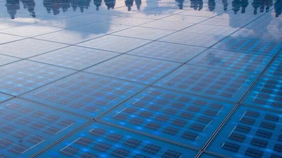 A large circular plaza with glowing blue glass floor panels and people standing around the edge at dusk.