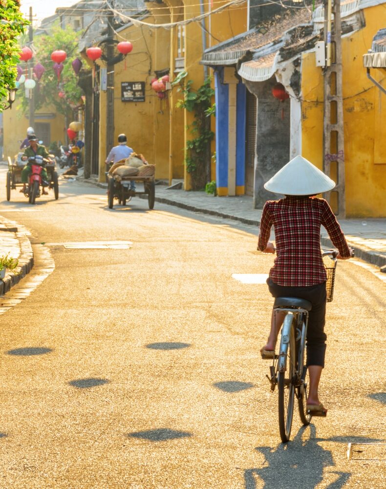 Hoi An (Hoian), Vietnam – April 12, 2018: Vietnamese woman in traditional bamboo hat bicycling along Hoi An Ancient Town. Awesome view of old street decorated with colorful silk lanterns at sunrise.