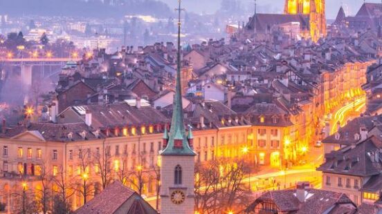 An elevated view of Bern Old Town at twilight, with illuminated spires and buildings in warm evening light.