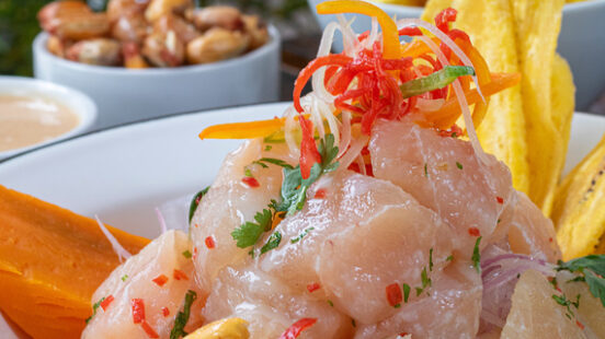 A white bowl of fresh fish ceviche topped with peppers and cilantro, served with toasted corn and plantain chips.