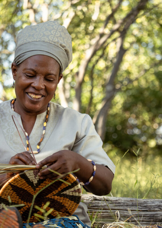 A smiling woman wearing traditional Botswanan clothing and headwrap weaving
