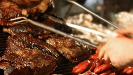 Multiple large steaks and sausages being grilled on a professional barbecue with metal tongs visible.