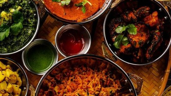 Multiple bowls of Indian food like biryani and curry served with flatbread and dipping sauces on a woven tray.
