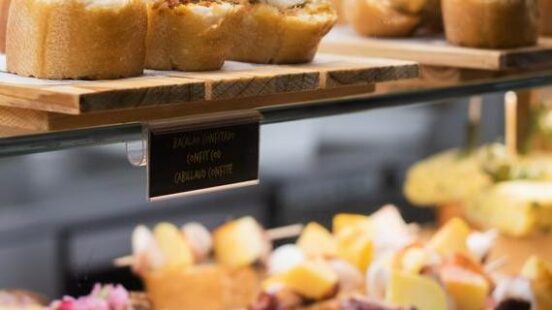 Assorted pinchos featuring fish and meat toppings on bread slices arranged on shelves in a food display.
