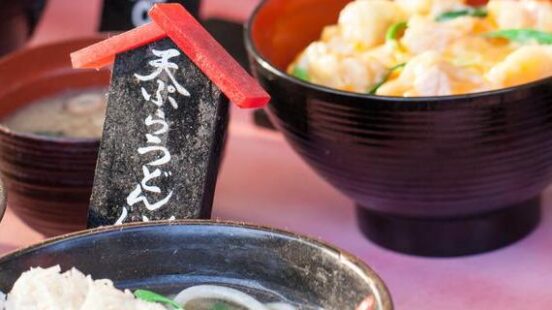 Close-up of Japanese food bowls including tempura noodle soup with decorative signs in the background.