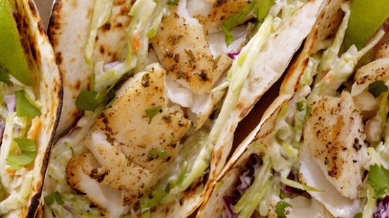 Three fish tacos topped with slaw and cilantro sitting on brown parchment paper with lime slices.