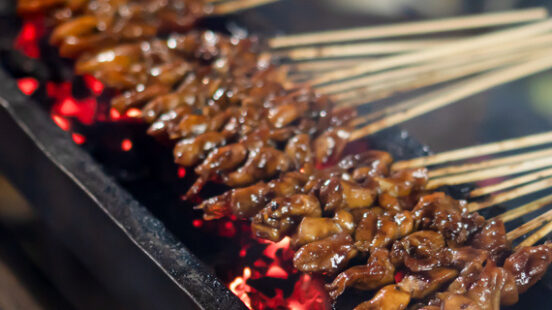 Dozens of meat satay skewers cooking over a long narrow charcoal grill with glowing red embers.