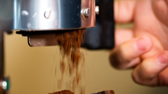 Close-up of ground coffee falling from a metal grinder into a portafilter held by a personx27;s hands.